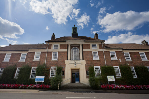 Бетлемская королевская больница — англ. Bethlem Royal Hospital), первоначальное название — госпиталь святой Марии Вифлеемской, психиатрическая больница в Лондоне. Здание с краснокирпичным фасадом и белыми оконными рамами, украшенное зеленью и цветами, с центральным входом, ведущим по лестнице. Над входом находится арочное окно, а на крыше возвышается небольшая башенка с часами. На фоне голубое небо с белыми облаками.