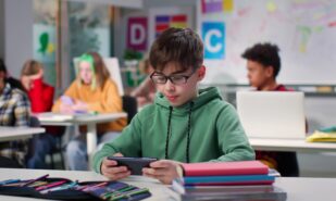 Teenage boy watching video on smartphone sitting at desk in class. Schoolboy using cellphone in classroom at school