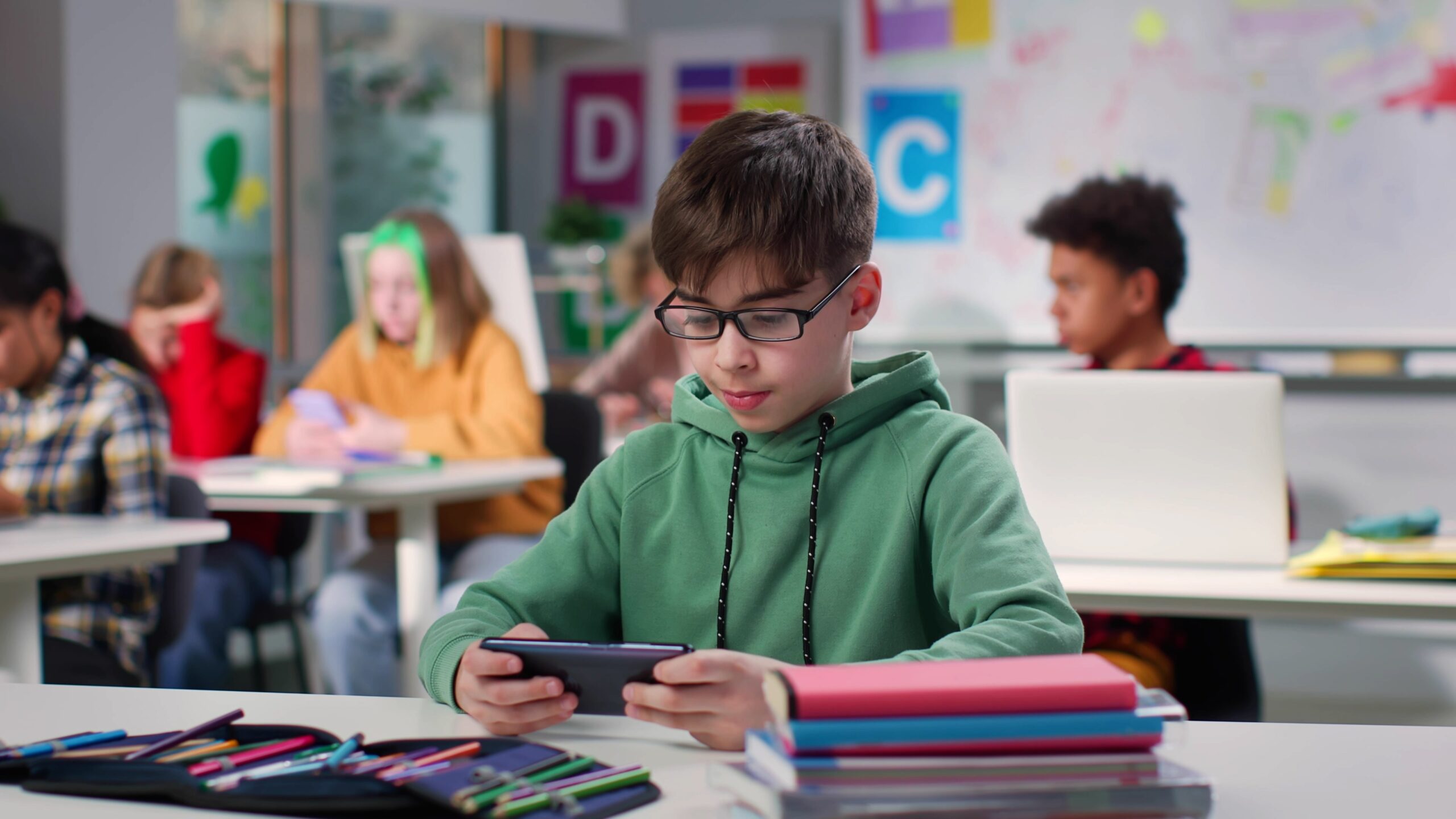 Teenage boy watching video on smartphone sitting at desk in class. Schoolboy using cellphone in classroom at school