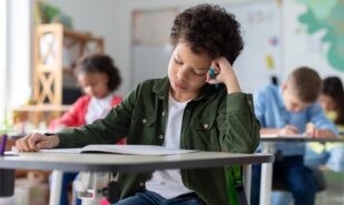 Tired and bored boy sitting at desk in classroom at school, writing in notebook and thinking, resting head on hand