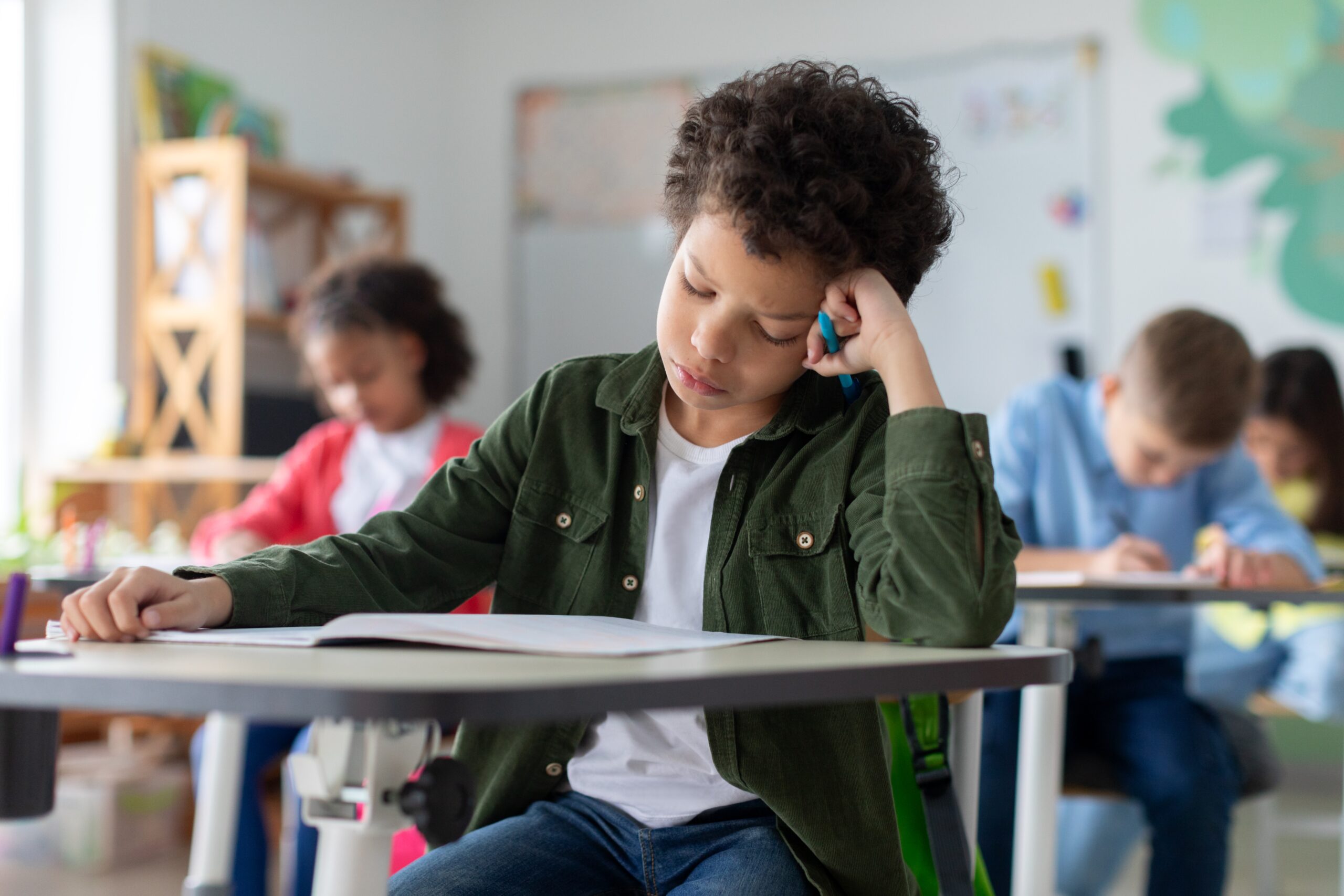 Tired and bored boy sitting at desk in classroom at school, writing in notebook and thinking, resting head on hand