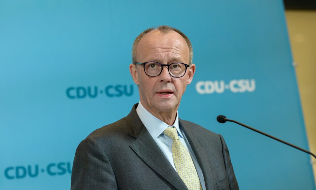 Friedrich Merz speaking at a meeting in Berlin in a microphone in front of a blue background wall