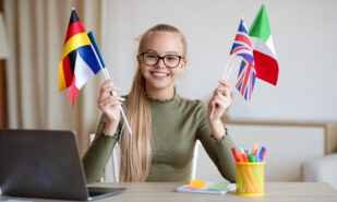 Cheerful blonde teen girl with glasses sitting at workdesk with international flags, home interior, using laptop, studying foreign languages. International education, online language courses