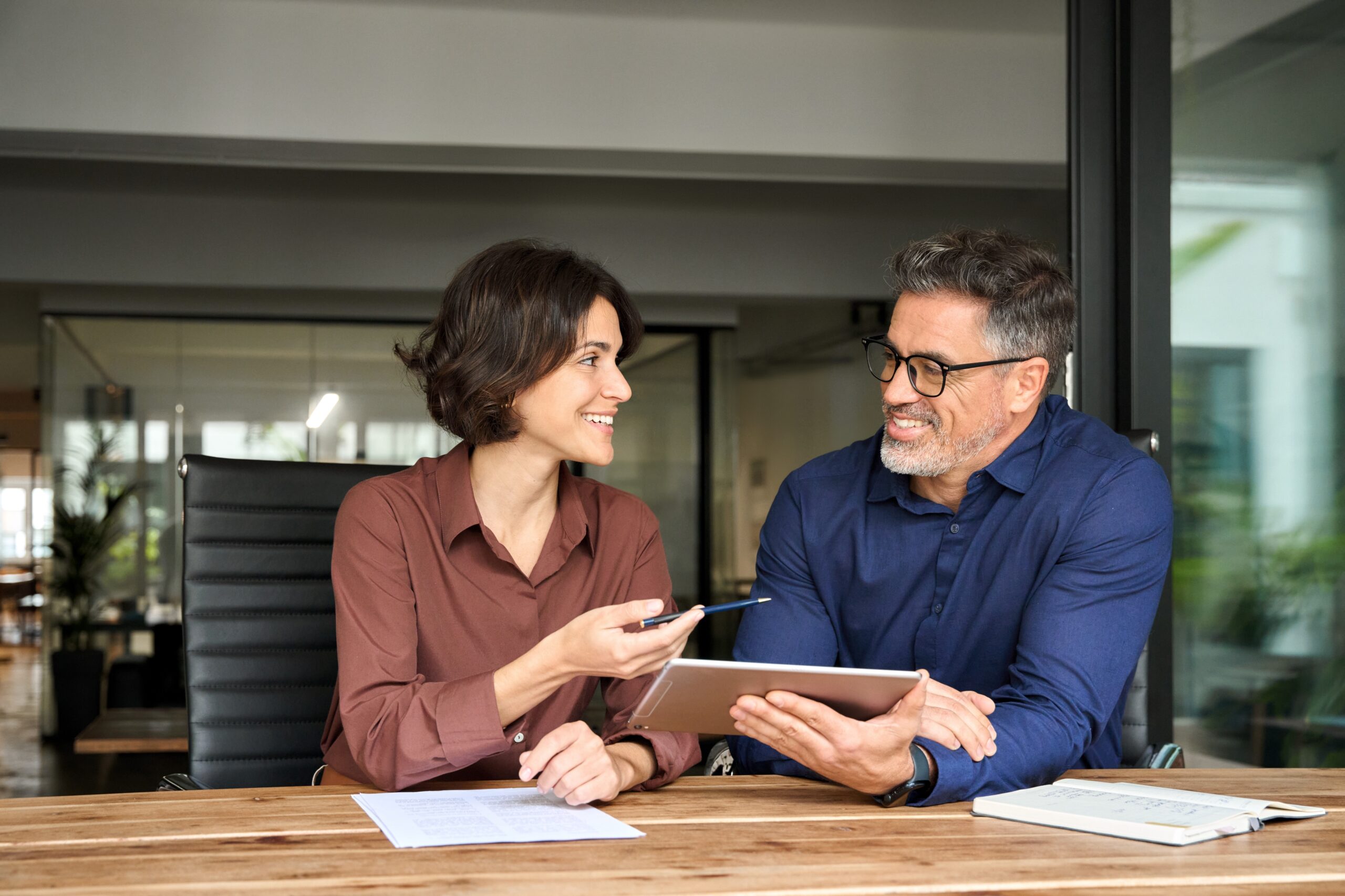 Two happy busy business colleagues having conversation at corporate meeting. Business team people having discussion planning sales financial strategy in company using tablet working together in office