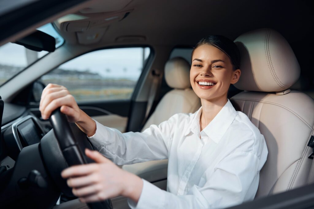 Smiling woman driving a car in a parking lot, hands on the steering wheel, joyful expression, safe driving, sunlight through windshield