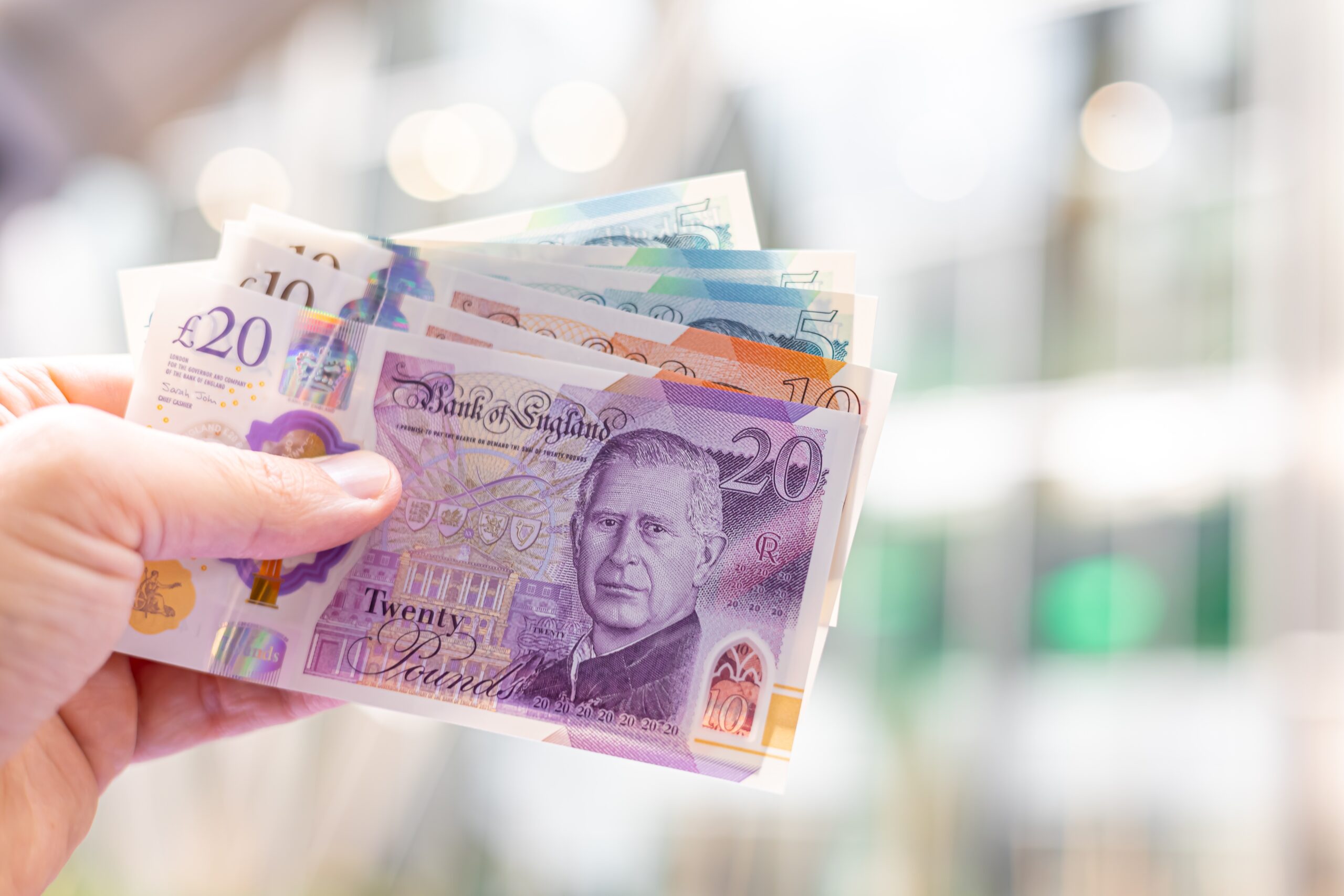 Close-up of a hand holding British pound banknotes (£5, £10, £20), featuring King Charles III. Useful for finance, banking, currency exchange, and economic themes.
