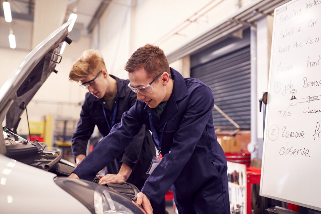 Two Male Students Studying For Auto Mechanic Apprenticeship At College Working On Car Engine