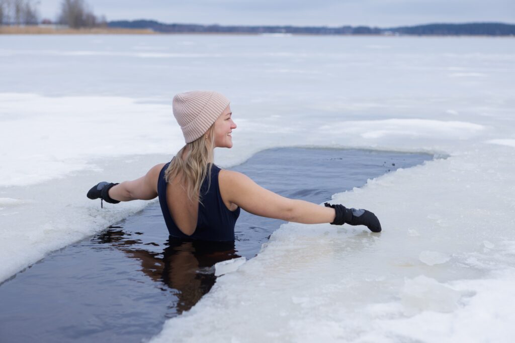 Winter swimming. Woman in frozen lake ice hole. Swimmers wellness in icy water. How to swim in cold water. Beautiful young female in zen meditation. Gray hat and gloves swimming clothes. Nature lake