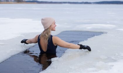 Winter swimming. Woman in frozen lake ice hole. Swimmers wellness in icy water. How to swim in cold water. Beautiful young female in zen meditation. Gray hat and gloves swimming clothes. Nature lake