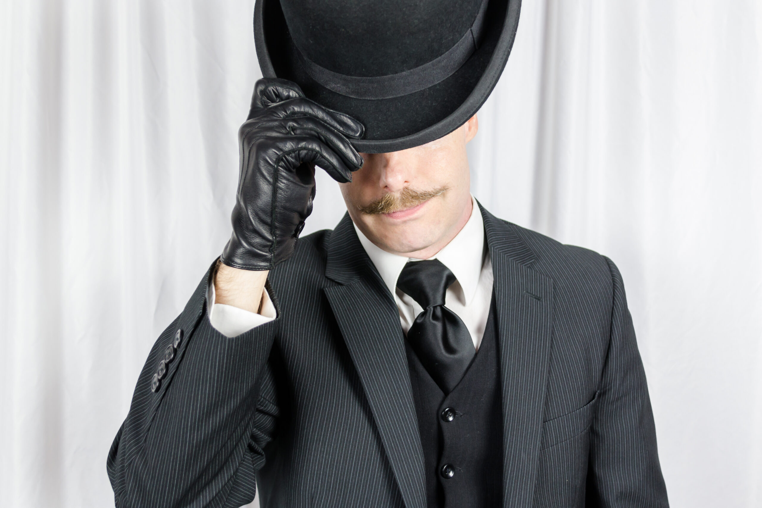 Portrait of British Gentleman in Dark Suit and Leather Gloves Politely Doffing Bowler Hat. Suave Spy Hero with Jaunty Moustache.