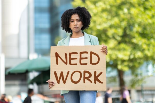 Unemployed pretty young black woman wearing casual outfit standing on street, holding Need Work placard, upset unhappy african american lady attending unemployment strike