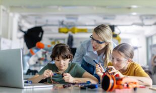 Female teacher helping to girls working on small robot, building robotic car in after-school robotics club. Children learning robotics in Elementary school. Girls in science.